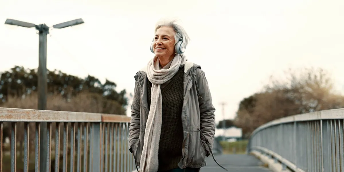 senior woman, headphones and music outdoor, walking and wellness with audio streaming and energy podcast, listen to radio and sound with female person on city bridge, exercise and travel with tech