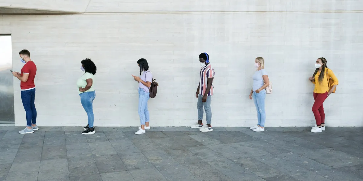 multiracial people standing in a queue and waiting - young people with social distancing and wearing protective face masks - concept of the new normality and social distancing