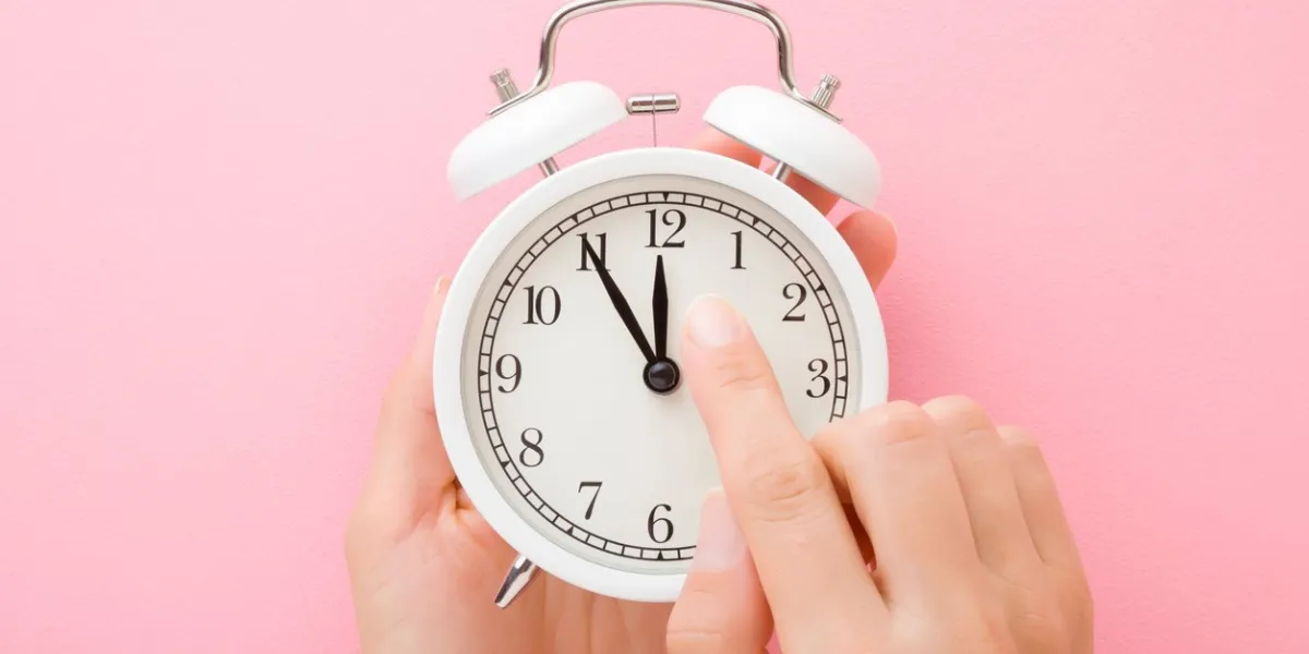 young woman hands holding white alarm clock finger pointing to arrow of twelve o'clock light pink table background pastel color time change concept closeup top down view