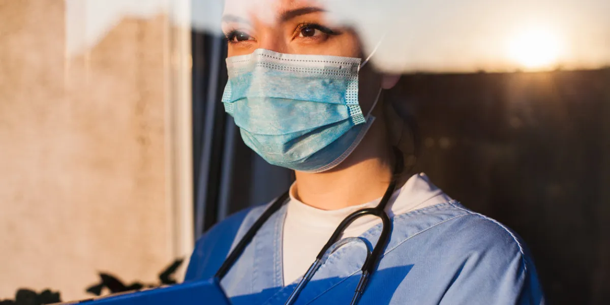 young sad female caucasian nhs ems doctor carer looking through icu window, fear uncertainty in eyes, wearing face mask gazing at sun ,hope and faith in overcoming coronavirus covid-19 pandemic crisis