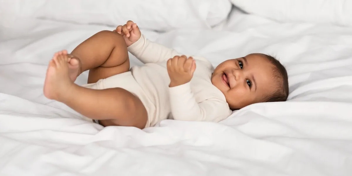 in the morning portrait of adorable smiling black infant lying on the back in bed on white bedsheet at home positive healthy laughing afro child wearing bodysuit, looking at camera happy childhood