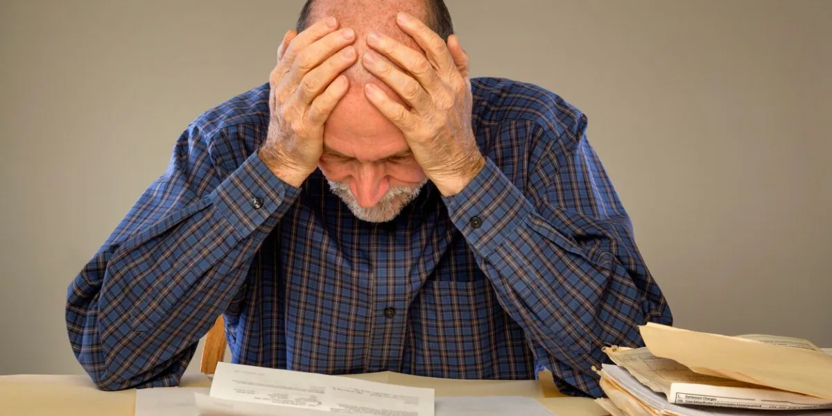 a senior adult man sitting at a table or desk stacked with papers and envelopes looking down with his hands on his head