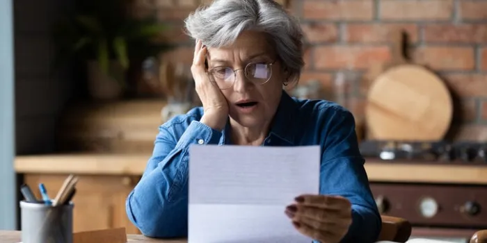 unhappy mature wearing glasses reading bad news in letter, stressed grey haired female with open mouth looking at paper sheet, sitting at desk, shocked by negative message in correspondence