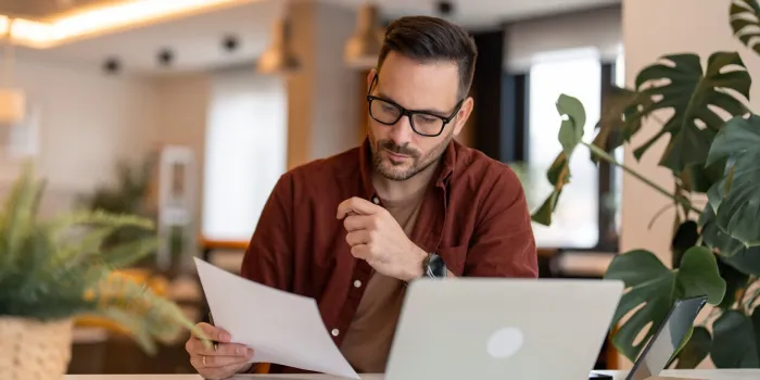 serious millennial man using laptop sitting at the table in a home office, focused guy in casual clothing looking at the paper, communicating online, writing emails, distantly working or studying on computer at home