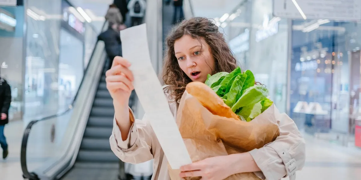 a woman in shock looks into a paper check from a supermarket in a shopping center against the background of an escalator and holds a package with fresh products, price increase