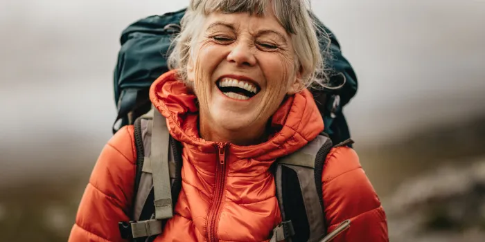 close up of a senior woman wearing jacket and backpack laughing with eyes closed portrait of a woman hiker laughing during her trekking
