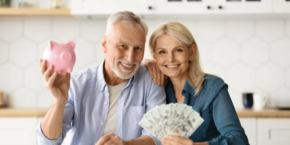 investments concept smiling senior couple holding piggybank and dollar cash in hands at home, happy elderly spouses saving money, advertising private pension plan, posing in kitchen interior