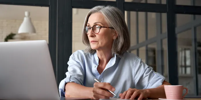 serious mature older adult woman watching training webinar on laptop working from home or in office 60s middle aged businesswoman taking notes while using computer technology sitting at table