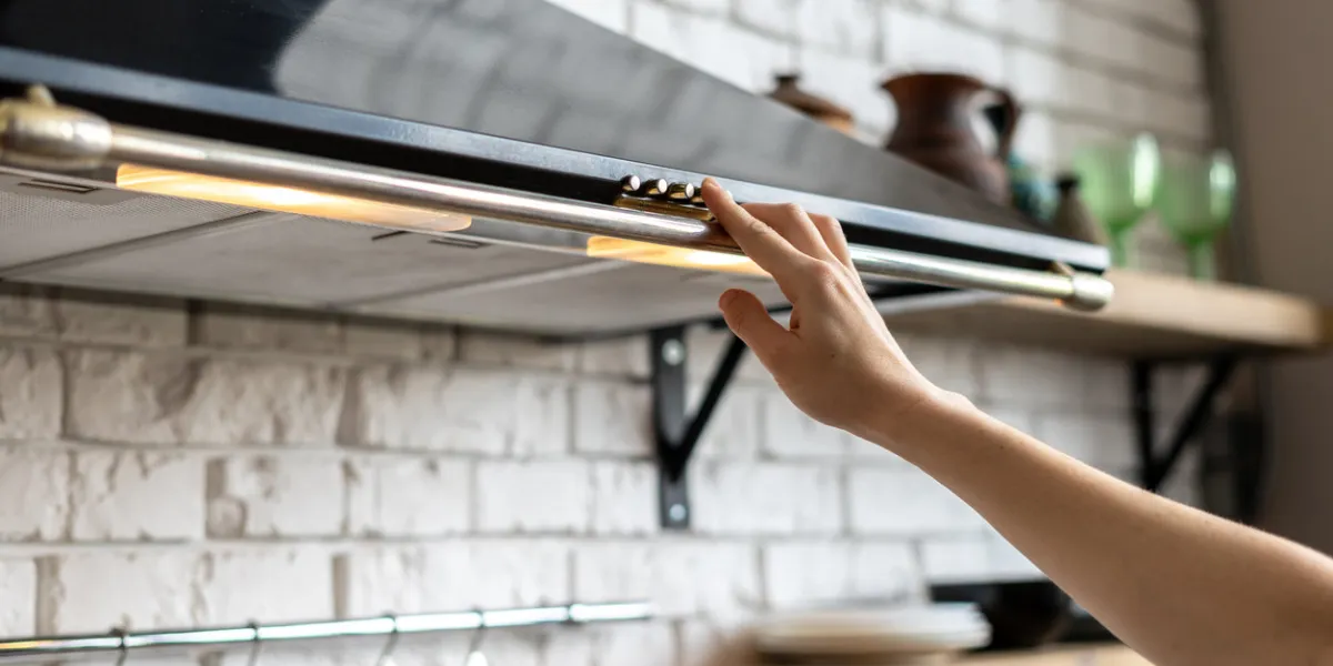 cropped view of woman hand select mode on cooking hood, standing near kitchen appliance in contemporary interior with brick wall and decor on shelves at blurred background