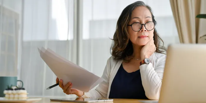concentrated asian middle aged female teacher or businesswoman in glasses sitting at desk using portable computer and examining paperwork age and technology
