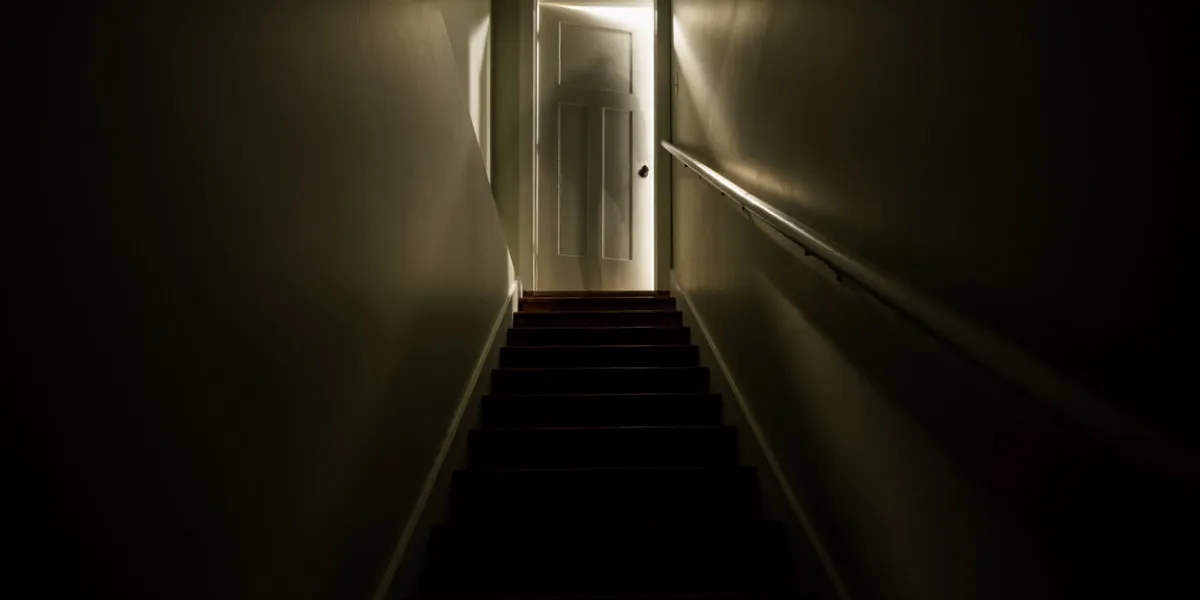 a dark stairwell illuminated by a slightly opened door at the top of the stairs shot with a long exposure to create the effect of a sillhouette of a ghost like figure at the top of the stairwell