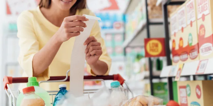 smiling woman doing grocery shopping at the supermarket and checking a long receipt, she is leaning on the full cart