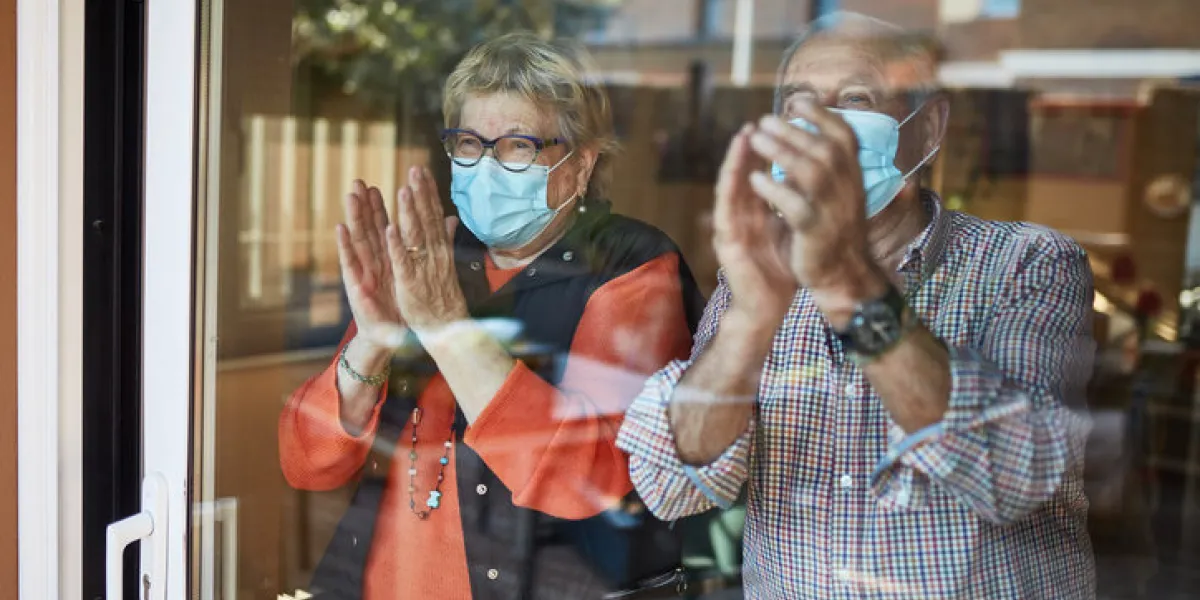 senior couple in self isolation looking through the window at home clapping they are showing their support for all the workers and helpers who are helping during the covid-19 outbreak