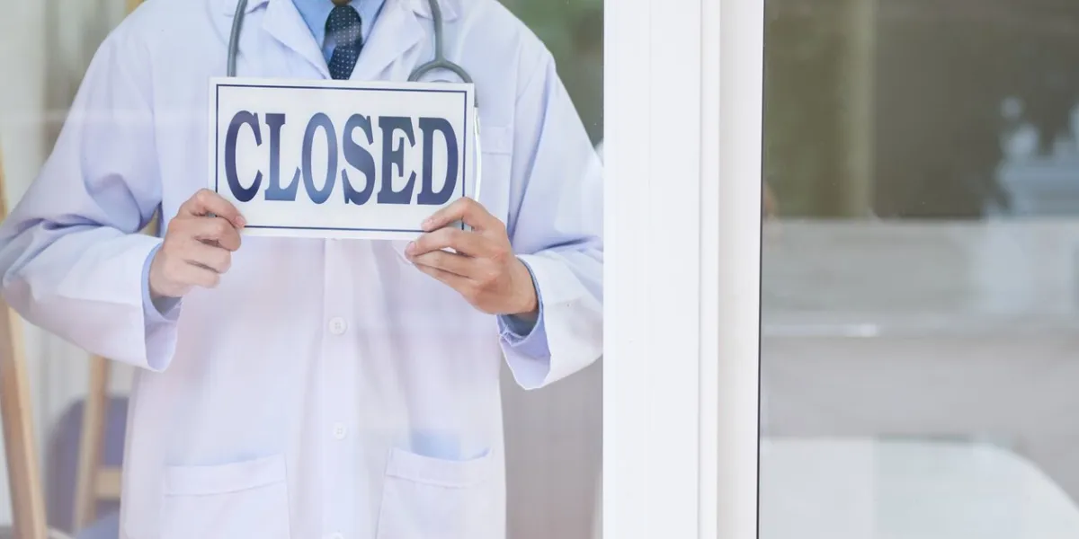 anonymous guy in medical uniform holding sign with closed writing while standing behind glass in office