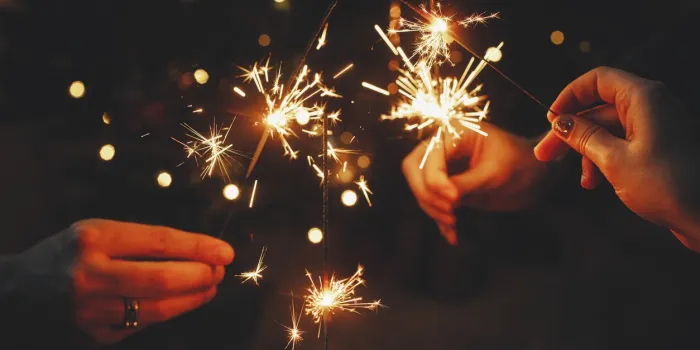 happy new year friends celebrating with burning sparklers in hands against christmas tree lights in dark room hands holding fireworks on background of stylish decorated illuminated tree moody