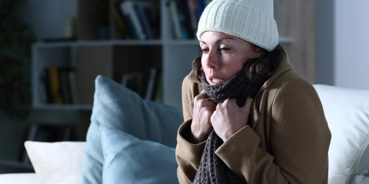 cold adult woman covered with clothes freezing sitting on the sofa at night at home