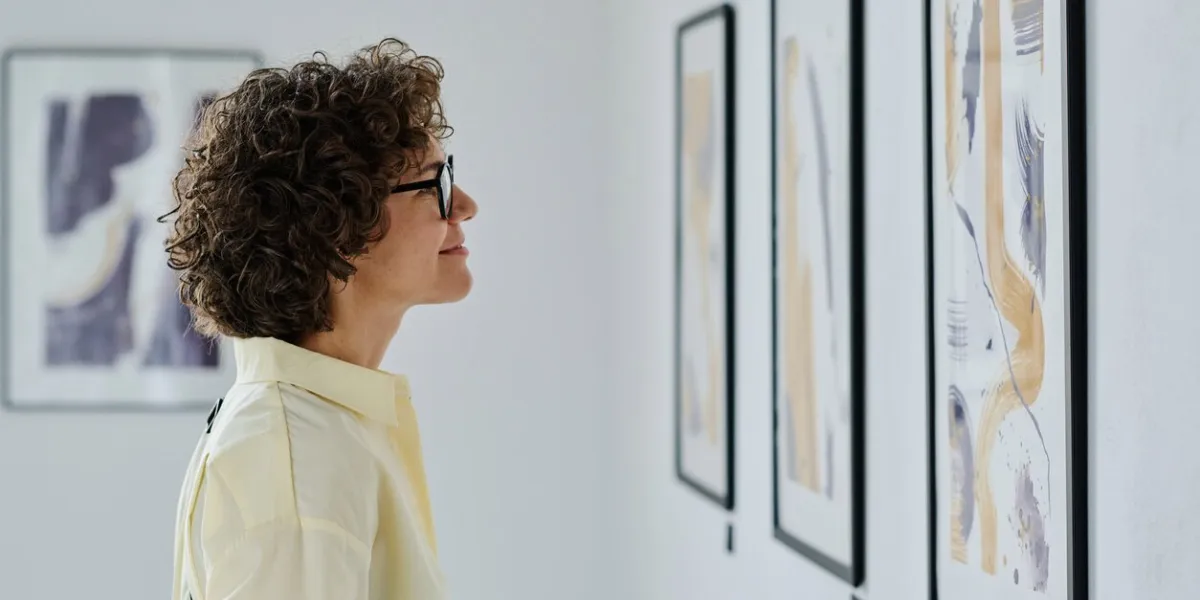 young woman in eyeglasses standing in front of pictures on the wall, examining the art and smiling