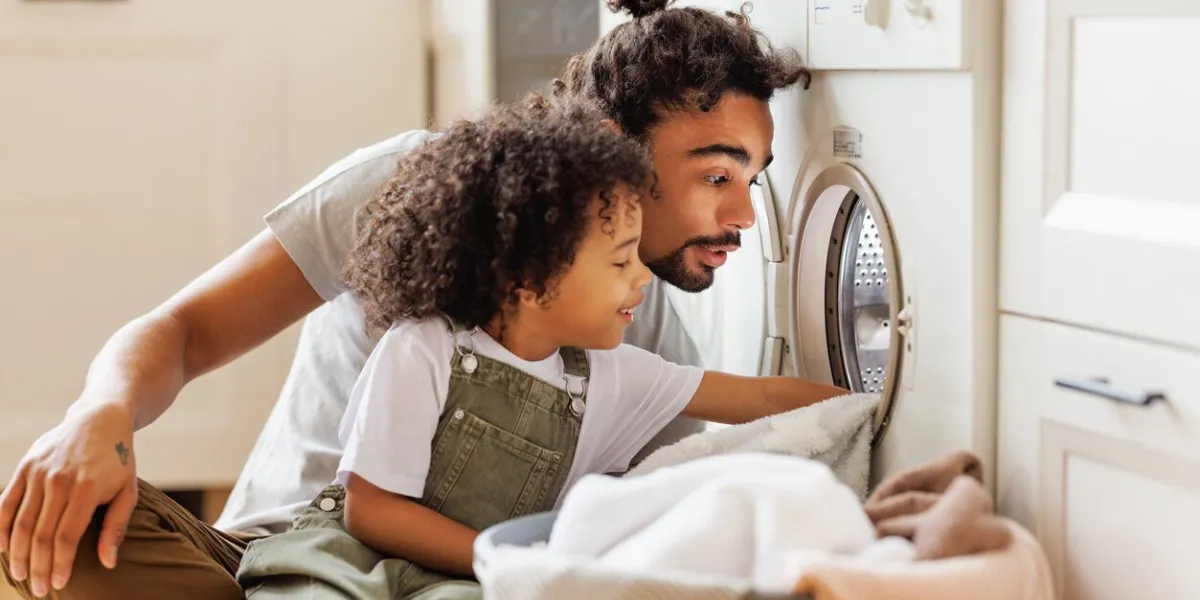 cheerful black kid boy sitting on dad's lap and helping father at linen in basket while doing laundry near washing machine in flight kitchen in weekend at home