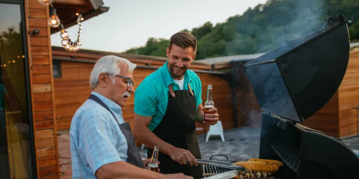 pensioner and his son wearing aprons, holding bottles of beer and roasting meat on barbecue in the backyard