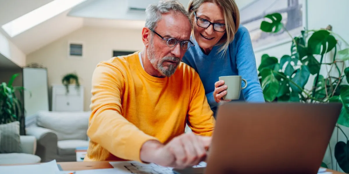 happy senior couple laughing and bonding while using laptop at home smiling elderly husband and wife having fun satisfied with buying insurance, paying bills online man showing something on laptop