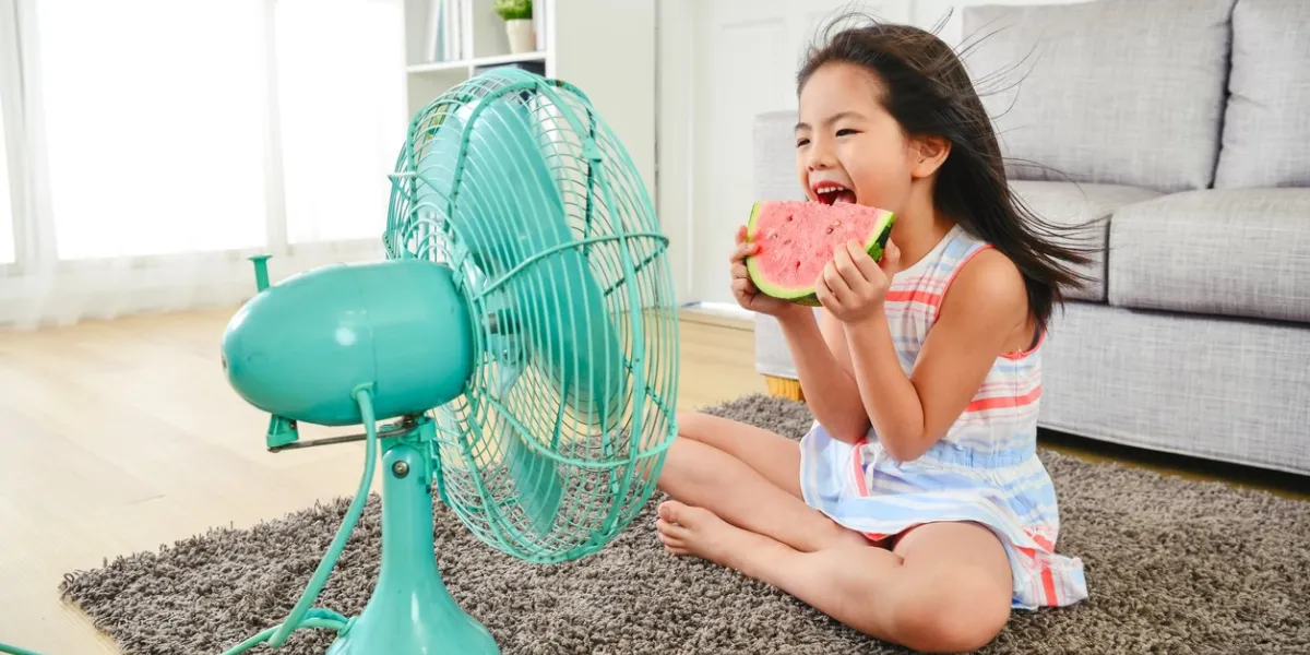 child eating watermelon with two hands sitting in front of the electric fan taking a big bite delightfully