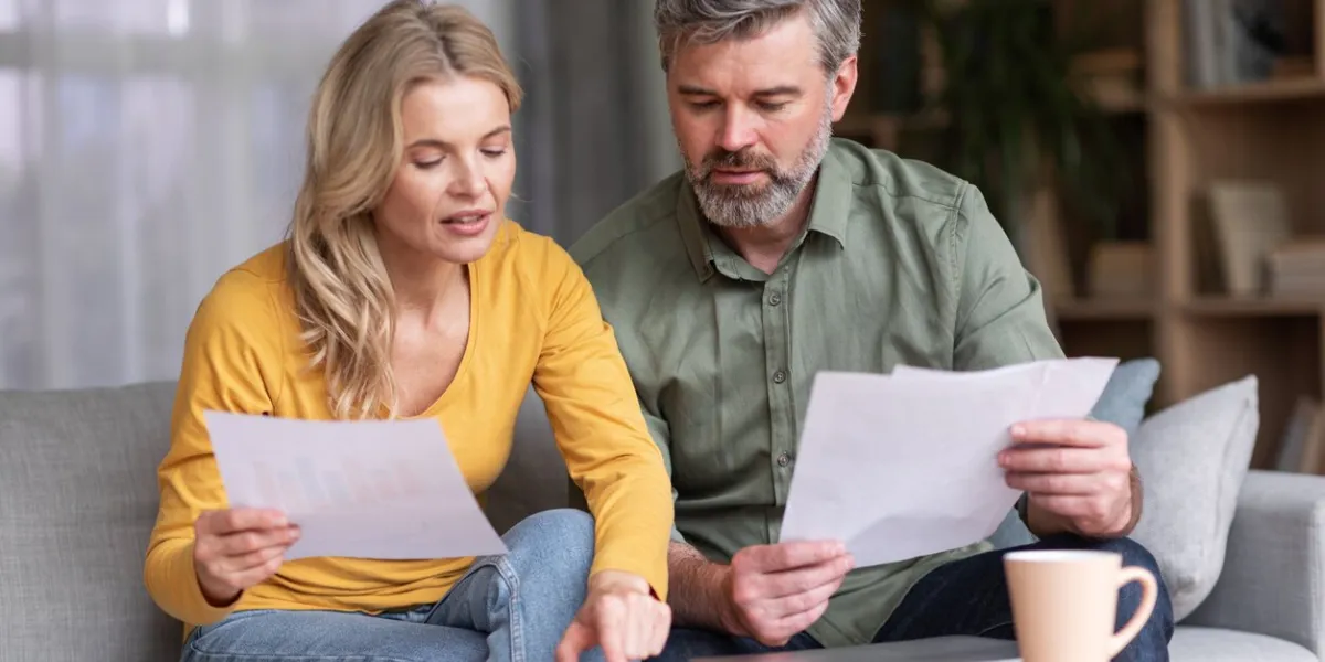 financial literacy busy middle aged husband and wife calculating family budget together while sitting on couch in living room interior, spouses checking loan documents and using calculator