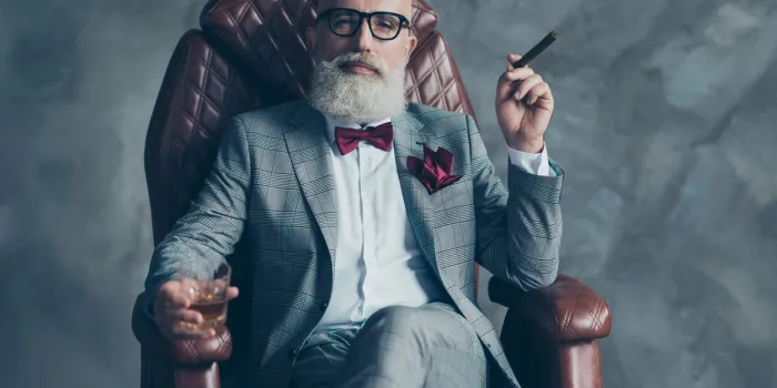 cool man in glasses, hold cigarette, glass with brandy, in formal wear, tux with red bowtie and pocket square, sit in leather chair over gray background, looking to the camera, shares, stock, money