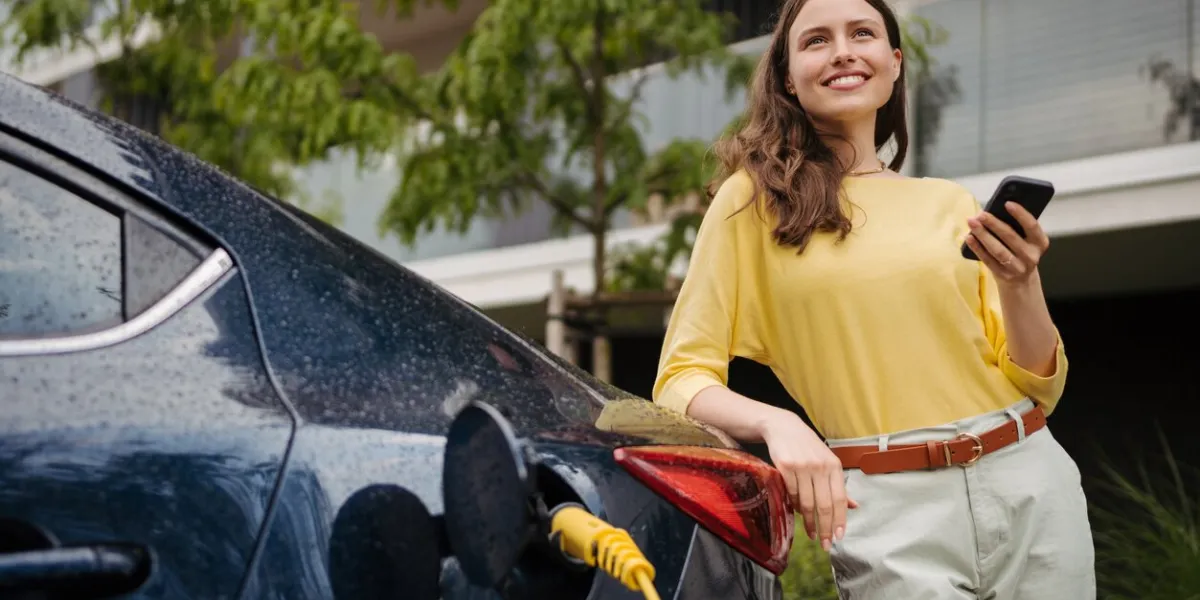 young woman with smartphone waiting while her electric car charging in home charging station, sustainable and economic transportation concept