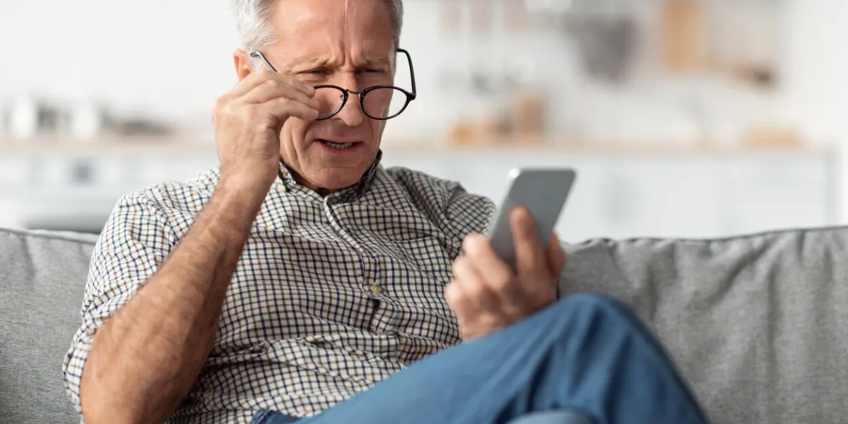poor eyesight senior man squinting eyes reading message on phone wearing eyeglasses having problems with vision sitting on couch at home ophtalmic issue, bad sight in older age concept