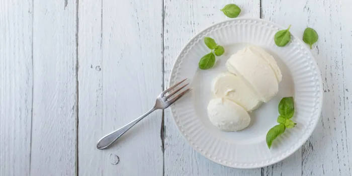 fresh cut whole mozzarella cheese served ready to eat with a fork and basil leaves on a white plate isolated on white wooden background side view, flat lay with copy space