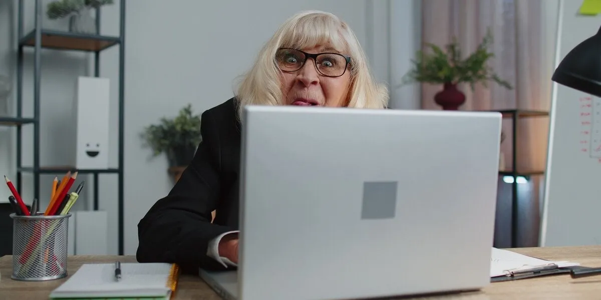 senior mature business woman lady hiding behind laptop computer, making funny face, fooling around, putting her tongue out remote working disrespecting someone at home office mature comic granny