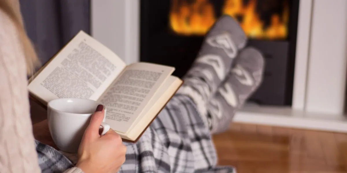 beautiful young woman in front of the fireplace reading book and warming feet on fire and legs are covered with blanket, in hand holds cup of hot tea winter and cold weather concept at home close up, selective focus