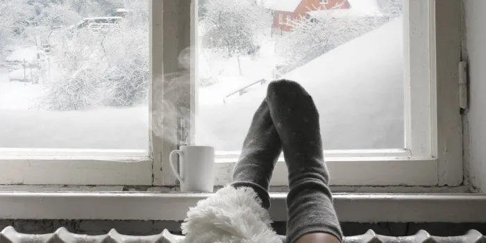 cozy winter still life  woman legs in warm woolen socks under shaggy blanket and mug of hot beverage on old windowsill against snow landscape from outside