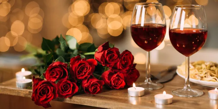 romantic dinner bouquet of flowers lying on the table, selective focus on bunch of roses, two glasses of red wine and candles on the wooden desk date concept, blurred background, banner, copy space