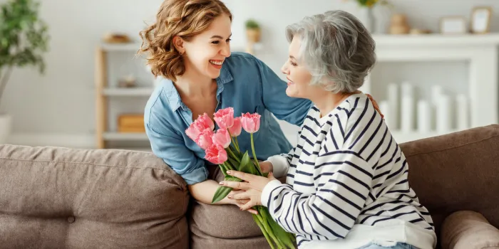 cheerful young female hugging happy aged mother and giving bouquet of tulips while congratulating on mother day at home