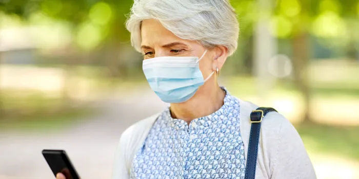 health, safety and pandemic concept - happy senior woman woman wearing medical mask for protection from virus with smartphone at summer park