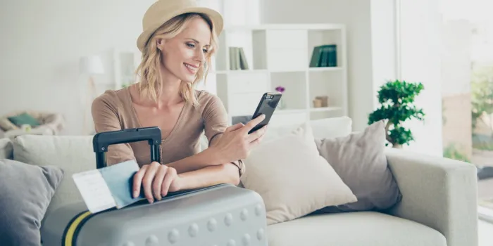 glad lady in casual denim jeans her curly wavy hair she sit on cozy sofa with luggage baggage light house interior hold document boarding pass in hand call taxi texting sms message on cellular gadget