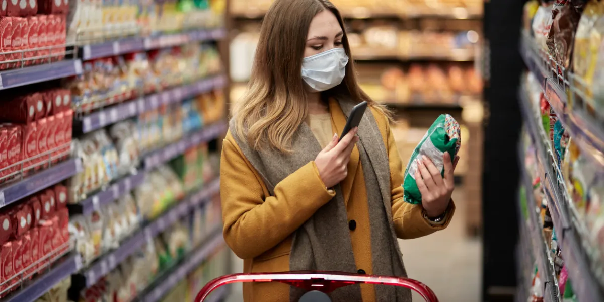 a young woman in a medical mask with a smartphone in her hand scans the product there is a large shopping cart nearby