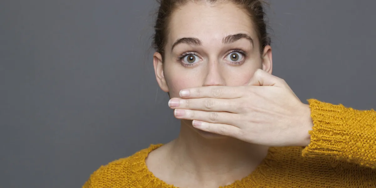 negative feelings concept - portrait of amazed beautiful 20s girl covering her mouth for gag metaphor,studio shot on gray background