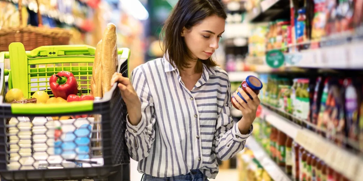 consumption and consumerism portrait of young woman with shopping cart in market buying groceries food taking products from shelves in store, holding glass jar of sauce, checking label or expiry date