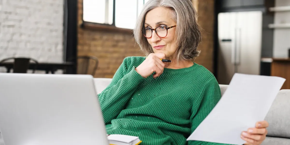 serious senior accountant attentively looking at laptop screen, examines the budget, documents of the company, on the living room, holding laptop on lap, working online, prepares an annual report