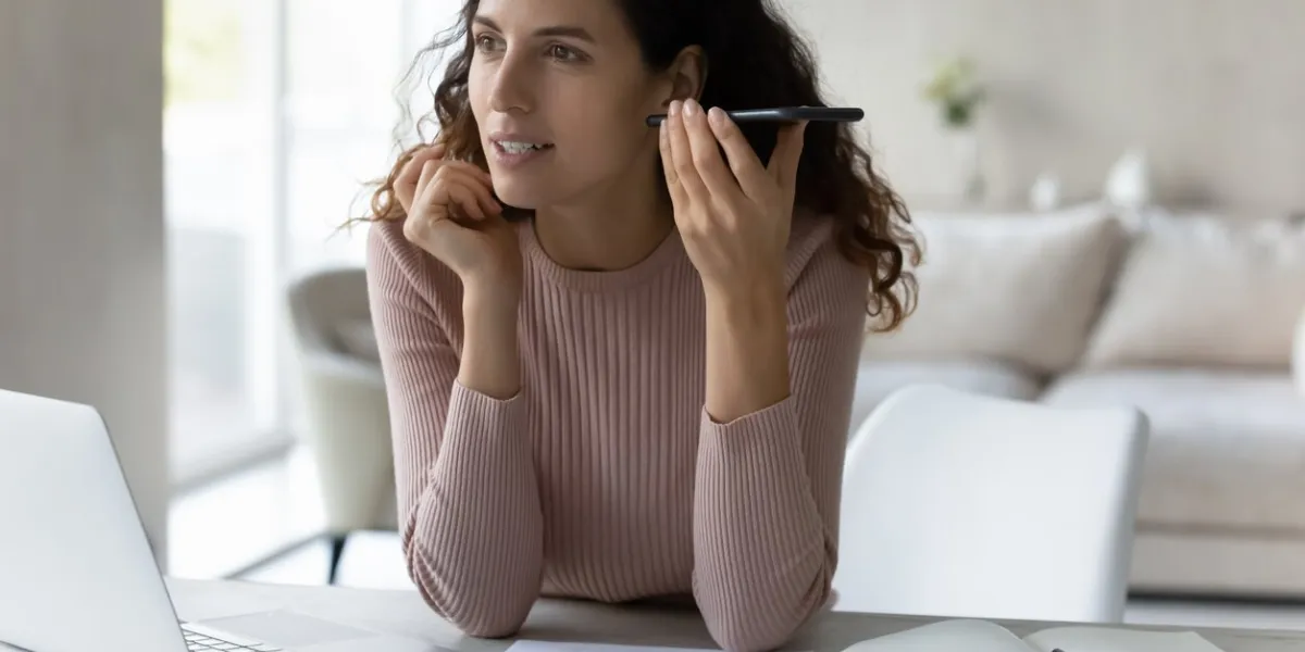 concentrated young hispanic business woman listening audio message on smartphone, sitting at table with computer focused millennial female employee worker communicating distantly using mobile apps