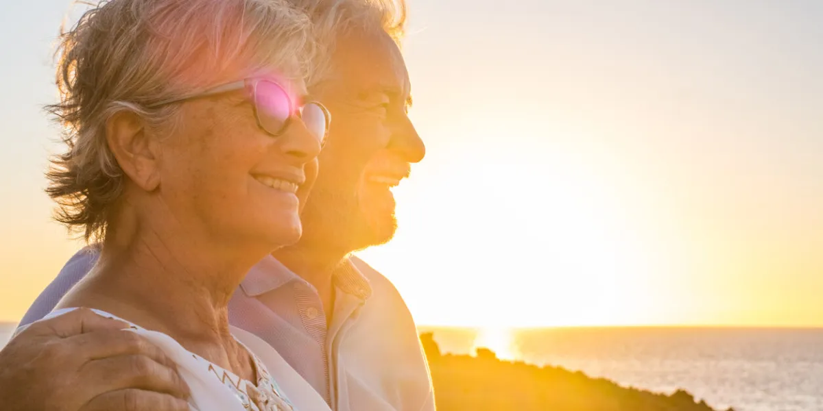 couple of two cute seniors together enjoying summer and having fun at the beach looking at the sea or ocean with sunset - mature people having a good lifestyle