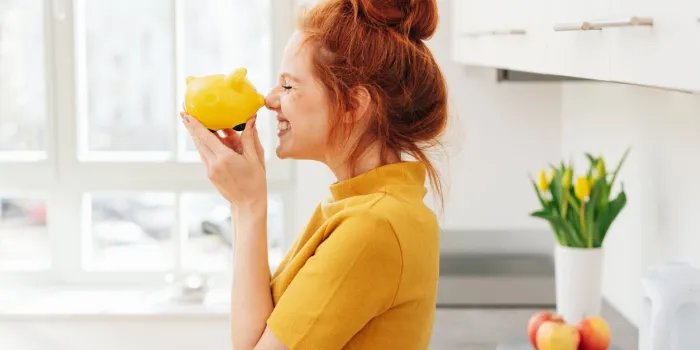 smiling red-haired woman playing with yellow piggy bank in her hands, viewed from the side in bright room interior money savings concept