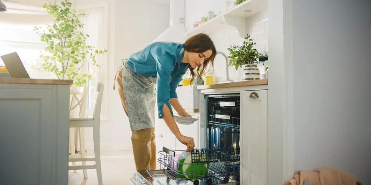 beautiful female is loading dirty plates into a dishwasher machine in a bright sunny kitchen girl in wearing an apron young housewife uses modern appliance to keep the home clean