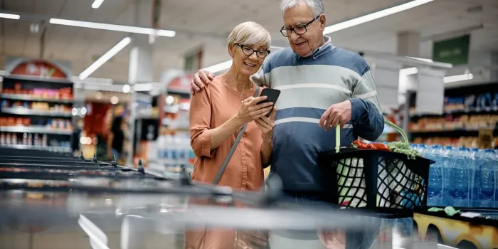 happy senior woman and her husband using app on mobile phone while shopping in supermarket