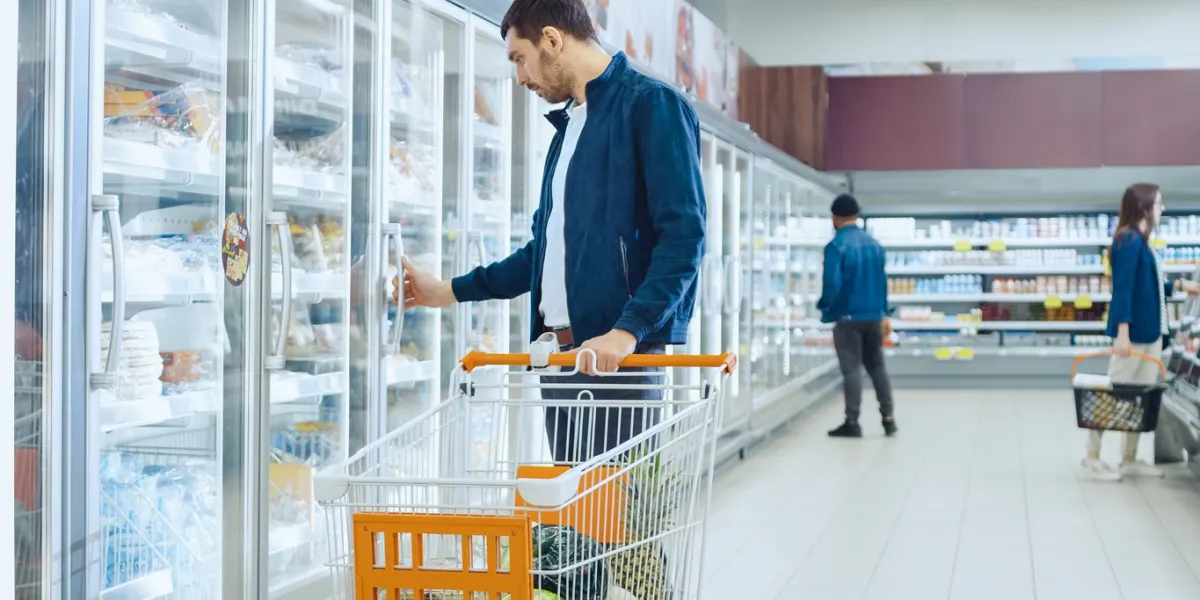 at the supermarket  handsome man pushes shopping card and browses for products in the frozen goods section man looks into glass door fridge, looking for dairy products other customer shopping in the background