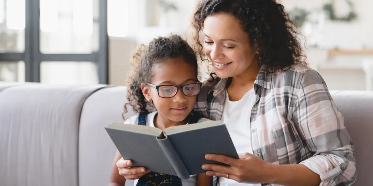 african-american mother mom reading fairy tales book novel with daughter girl, learning together, helping assisting with homework for school female tutor teaches schoolgirl at home homeschooling