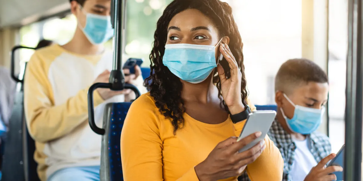 portrait of happy smiling black female in medical face mask traveling on public transit, listening to music touching wireless earphones and using cellphone, looking away at window, sitting inside
