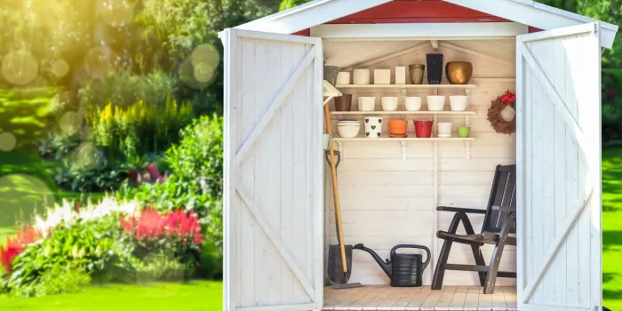 garden shed filled with gardening tools shovels, rake, pots, water pitcher in storage hut green sunny garden in the background